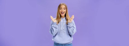 Photo for Portrait of amused excited and entertained happy charming woman with fair hair in trendy hoodie clasping hands from delight and surprise smiling broadly joyfully posing against purple background - Royalty Free Image