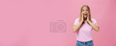 Photo for Delighted and amazed happy girlfriend in t-shirt and jeans smiling broadly with opened mouth amused touching cheeks surprised and satisfied reacting to positive surprise over pink background. Copy - Royalty Free Image