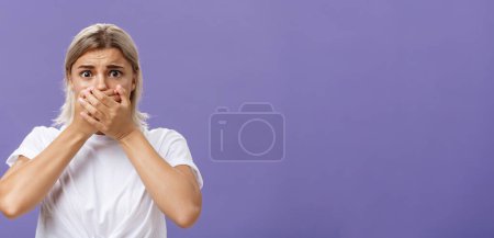 Photo for Waist-up shot of shocked nervous and scared young woman witnessing terrible crime covering mouth with both hands not to scream frowning staring frightened at camera over purple background. Emotions - Royalty Free Image