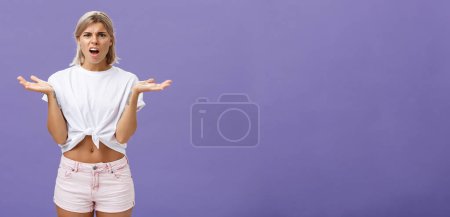 Foto de Estilo de vida. Perpleja y decepcionante estudiante rubia guapa en camiseta blanca y pantalones cortos rosados frunciendo el ceño encogiéndose de hombros con las manos extendidas diciendo qué demonios sobre la pared púrpura. - Imagen libre de derechos