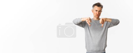 Photo for Skeptical and disappointed middle-aged man, grimacing unamused and showing thumbs-down, dislike something bad, standing over white background. - Royalty Free Image