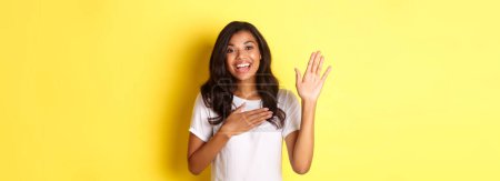 Téléchargez les photos : Portrait de fille afro-américaine mignonne et sincère, faisant une promesse, tenant la main levée et un autre sur le cœur, jurant de dire la vérité, debout sur fond jaune. - en image libre de droit