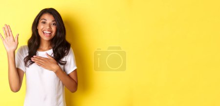 Téléchargez les photos : Portrait de fille afro-américaine mignonne et sincère, faisant une promesse, tenant la main levée et un autre sur le cœur, jurant de dire la vérité, debout sur fond jaune. - en image libre de droit