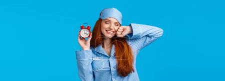 Photo for Girl slept well, wake up energized, sleepy stretching with closed eyes and lovely smile, holding red cute clock, set up alarm to be work in time, wearing pyjama and sleep mask, blue background. - Royalty Free Image