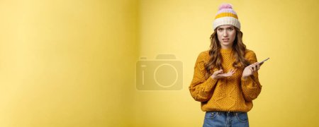 Photo for Confused cringing annoyed young woman shocked how friend talked her frowning raise hand dismay look frustrated camera holding smartphone finish video call unpleasant note, yellow background. - Royalty Free Image