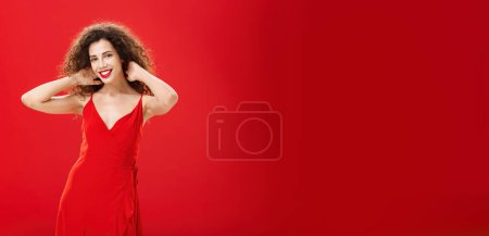 Téléchargez les photos : Élégant douce fille caucasienne avec coiffure bouclée et soirée maquillage touches brins de cheveux vérification coupe de cheveux et souriant largement à la caméra posant dans une nouvelle robe rouge élégante sur fond de studio - en image libre de droit
