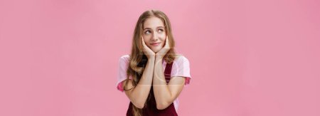 Photo for Girl wonders if she have sweets in fridge. Portrait of charming dreamy young woman in trendy outfit leaning head on palms gazing at upper left corner with tender smile daydreaming, feeling nostalgic - Royalty Free Image