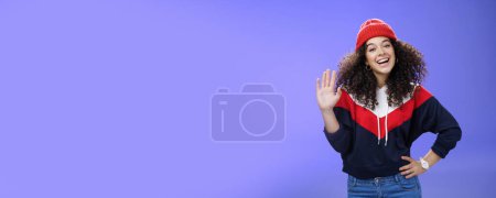 Photo for Hey nice to meet you. Charming female ski couch in cute red beanie with curly hair waving hello with palm and smiling broadly greeting newbies as teaching winter sports over blue background. Copy - Royalty Free Image