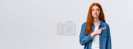 Photo for Awkward and nervous cute timid redhead teenage girl having fear or embarrassing thing say, pointing looking right anxious, biting lip and frowning, confessing in crime, feel guilty. - Royalty Free Image