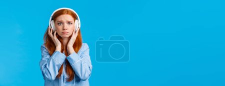 Photo for Girl about cry from emotional song. Upset redhead girlfriend suffering breakup, sobbing listening music in large white headphones, standing blue background in nightwear, unhappy. - Royalty Free Image