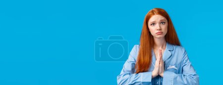 Photo for Please I am begging you. Silly and uneasy redhead caucasian girl in sleepwear, asking help, pleading forgiveness or need something, tilt head making sad face, standing blue background. - Royalty Free Image