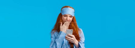 Photo for Gadgets addication, lifestyle and beauty concept. Lovely redhead girl going sleep and scrolling feed smartphone, yawning squinting feeling tired, wearing pyjama and sleep mask, blue background. - Royalty Free Image