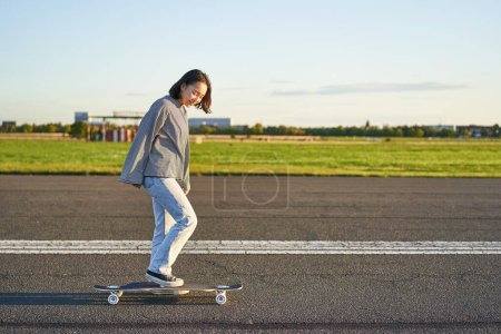 Téléchargez les photos : Belle fille patineuse asiatique chevauchant son longboard sur route vide ensoleillée. Jeune femme appréciant son tour de patin souriant et riant. - en image libre de droit