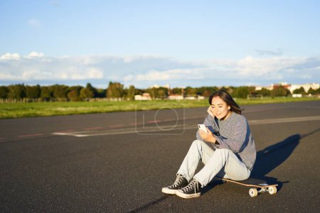 Téléchargez les photos : Skater girl assis sur son skateboard sur la route, en utilisant un smartphone, bavarder sur l'application mobile. - en image libre de droit