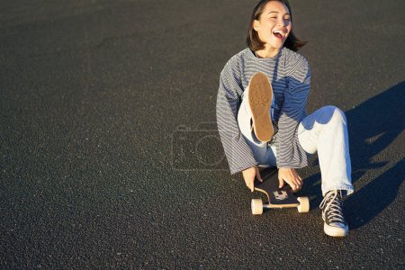 Téléchargez les photos : Portrait de belle fille asiatique patinant, assis sur son skateboard et souriant. Adolescent mignon avec longboard, patinage sur route. - en image libre de droit