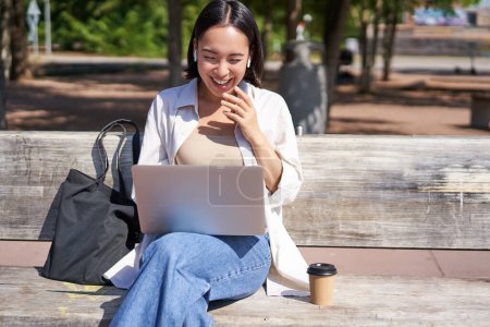 Photo for Young asian woman talking on video chat with laptop, sitting on bench, having online conference on remote. Copy space - Royalty Free Image