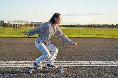 Téléchargez les photos : Joyeux patineur fille chevauchant son skateboard et s'amusant dans la rue vide. Femme souriante appréciant croisière sur route ensoleillée. - en image libre de droit
