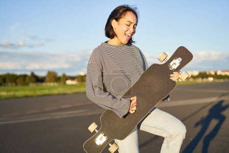 Téléchargez les photos : Belle asiatique adolescent fille jouer avec son longboard, tenant skateboard comme si jouer de la guitare, debout sur la route le jour ensoleillé. - en image libre de droit