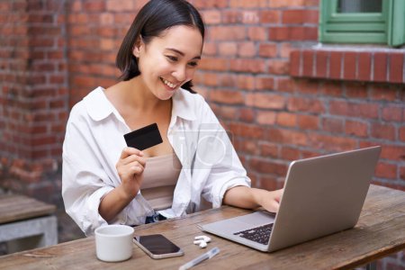 Photo for Smiling asian woman sitting with laptop, paying by credit card for online shopping, sending her bank account details, sitting in cafe. - Royalty Free Image