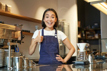 Portrait of smiling korean barista, girl at the counter, wears blue apron, works in coffee shop, shows thumbs up. People at work.