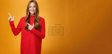 Photo for Assertive and helpful charming young female assitant in red knitted dress with ginger hair smiling friendly and pleasant as pointing at upper left corner, showing interesting thing over orange wall. - Royalty Free Image