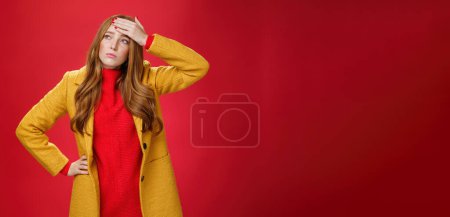 Photo for Girl having feaver touching forehead with palm looking displeased and bothered left as thinking she got cold standing in yellow coat, realising she sick or ill over red background, feeling tired. - Royalty Free Image