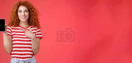 Téléchargez les photos : Et cette appli ? Redhead curly friendly cute girl help friend find oufit online store hold smartphone pointant l'écran du téléphone en souriant recommande largement le filtre photo sur les médias sociaux, fond rouge. - en image libre de droit