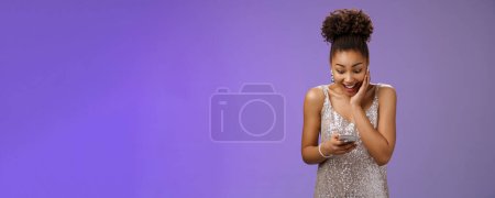 Photo for Surprised astonished young african american girl holding smartphone look display imressed super happy reading message touch cheek widen eyes gladly receive great news, standing blue background. - Royalty Free Image