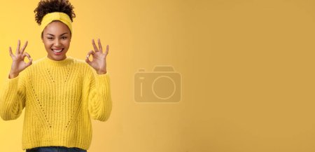 Foto de Cuéntalo hecho. Asegurada mujer afroamericana confiada en el show de diadema de suéter ok ok no te preocupes gesto sonriente plan seguro de sí mismo va bien, buenos resultados satisfechos, animando fondo amarillo. - Imagen libre de derechos