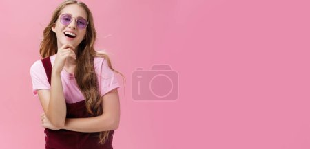 Photo for Self-confident good-looking pretty young lady with wavy hair floating on air raising head up having high self-esteem touching chin laughing and smiling wearing trendy sunglasses and dungarees. Body - Royalty Free Image