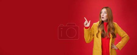 Téléchargez les photos : Mode de vie. Portrait de femme rousse surprise et choquée empathique en manteau jaune bouche ouverte étonnée et sans voix pointant et regardant le coin supérieur gauche concerné et secouée au-dessus du mur rouge. - en image libre de droit