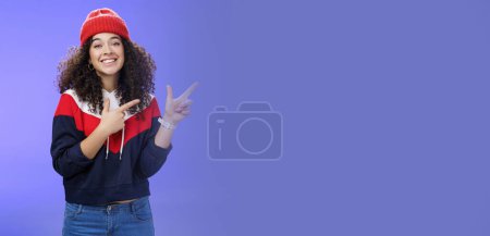 Photo for Cute female friend pointing at upper right corner with finger pistols as smiling broadly at camera wearing warm winter hat and sweatshirt skiing over blue background joyfully. Emotions, weather and - Royalty Free Image