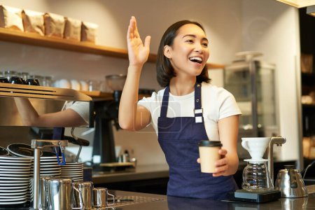 Foto de Amistosa chica barita sonriente saludando al cliente, sosteniendo su taza de café, orden de comida para llevar preparada, trabajando en la cafetería detrás del mostrador. - Imagen libre de derechos