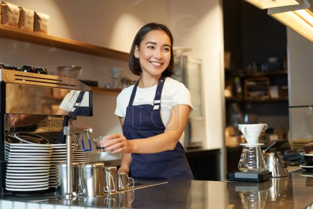 Smiling girl barista in cafe, preparing cappuccino in coffee machine, steaming milk, wearing uniform apron.