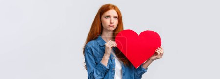 Photo for Unhapy brokenhearted cute redhead girl, sulking and looking up uneasy, distressed holding big red valentines day heart, was rejected, showed her feelings standing white background. - Royalty Free Image