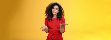 Photo for So waht, I confused. Questioned uncertain woman with curly hair in red dress shrugging looking clueless as holding hand cannot understand where order as checking mail box via device over yellow wall - Royalty Free Image
