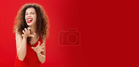 Téléchargez les photos : Roar va te manger chérie. Sensuelle et séduisante femme européenne ludique avec coiffure bouclée jouant dans la chambre à coucher avec amant en robe de soirée rouge faisant pattes d'animaux sortant de la langue et clin d'oeil. - en image libre de droit
