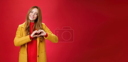 Photo for Love, romance and fall concept. Portrait of charming tender and gentle young redhead woman in yellow coat showing heart gesture making confession in sympathy, smiling cute at camera over red wall. - Royalty Free Image