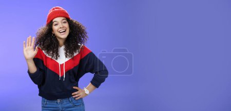 Photo for Hey nice to meet you. Charming female ski couch in cute red beanie with curly hair waving hello with palm and smiling broadly greeting newbies as teaching winter sports over blue background. Copy - Royalty Free Image