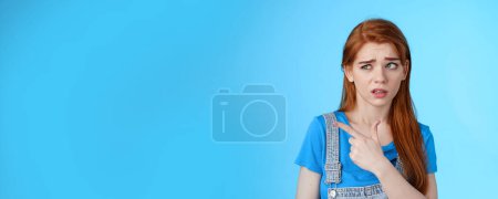 Photo for Stupified redhead hesitant girl show aversion, pointing looking left reluctant, full disbelief, stare with dismay uncertain about product, disliking strange weird guy standing blue background. - Royalty Free Image