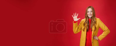 Téléchargez les photos : Fille donnant cinq étoiles à la boutique en ligne impressionnante, elle a acheté une tenue pour le temps d'automne montrant palmier levé souriant largement comme debout dans un élégant manteau jaune chaud sur fond rouge. Émotions et - en image libre de droit