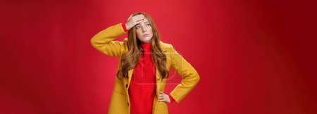 Photo for Girl having feaver touching forehead with palm looking displeased and bothered left as thinking she got cold standing in yellow coat, realising she sick or ill over red background, feeling tired. - Royalty Free Image
