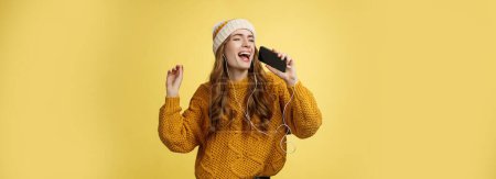 Photo for Carefree charming girl likes karaoke having fun listen favorite songs wearing wired earphones hold smartphone microphone singing out loud dancing enjoying spend time alone. Technology concept - Royalty Free Image