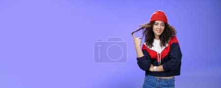 Photo for Stylish and snobbish arrogant curly-haired woman in warm beanie rolling curl on finger smirking and looking with contempt at camera, scorning person as being too cool over blue background. - Royalty Free Image