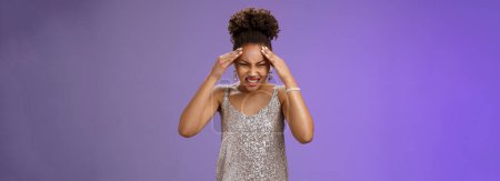 Photo for Pressured intense worried african-american woman in silver glittering dress grimacing cringing painful headache hold hand forehead suffer pain migraine, standing bothered blue background. - Royalty Free Image