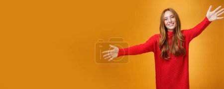 Foto de Ven y abrázame. Retrato de dulce hermana pelirroja femenina y linda en vestido de punto rojo extendiendo las manos hacia la cámara y sonriendo ampliamente dando una cálida bienvenida, queriendo abrazar sobre la pared naranja. - Imagen libre de derechos