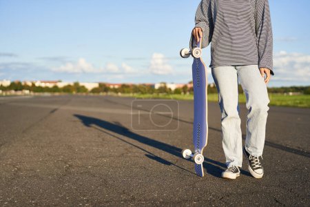 Téléchargez les photos : Coupé coup de corps de femme, tenant skateboard dans la main, marchant sur la route. Jeune femme porter son longboard, cruiser, patiner sur la route. - en image libre de droit