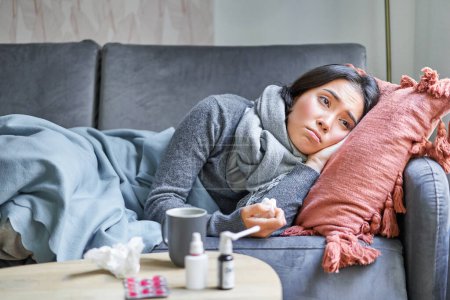 Photo for Sick sad korean woman lying on sofa, feeling unwell, catching cold, flu and temperature, looking upset, taking medication. - Royalty Free Image