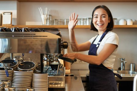 Photo for Enthusiastic asian barista, girl using coffee machine, waving at client, saying hello, greeting guests in her cafe. - Royalty Free Image
