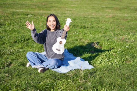 Téléchargez les photos : Musique et instruments. gai fille musicien asiatique femme tenir son ukulele joue dans parc et vagues main à vous dit bonjour à un ami. - en image libre de droit